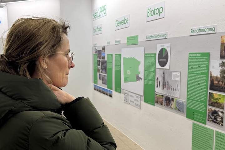 Frau mit braunen langen Haaren und Brille in grüner Winterjacke sieht sich Ausstellung über Biotope an. 